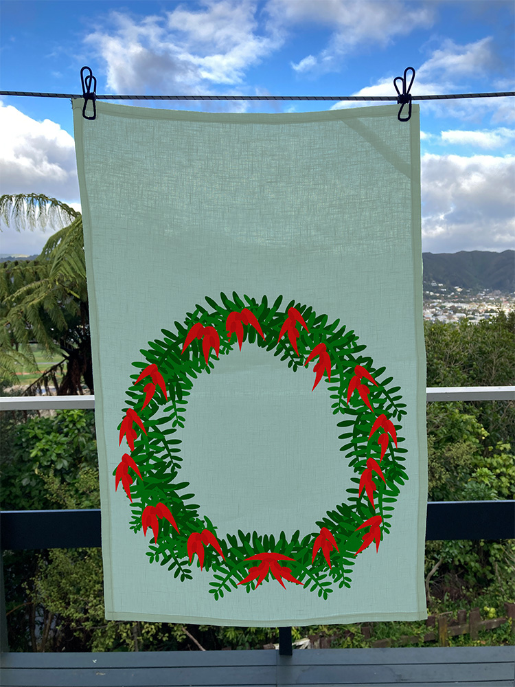 Wreath tea towel is printed with a large wreath design. The wreath is made up of very bright ngutukākā flowers and foliage on a pale green background. Here it is shown hanging on a line with tree canopy in the background.